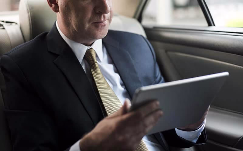 Book a black car for a business trip: executive in a suit working on a tablet in a luxury sedan.