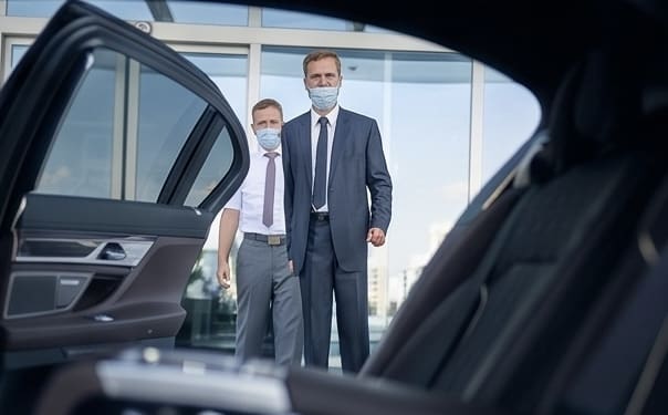 A professional chauffeur in a suit and protective face mask holding open the door of a black luxury sedan for an executive client in St. Louis.